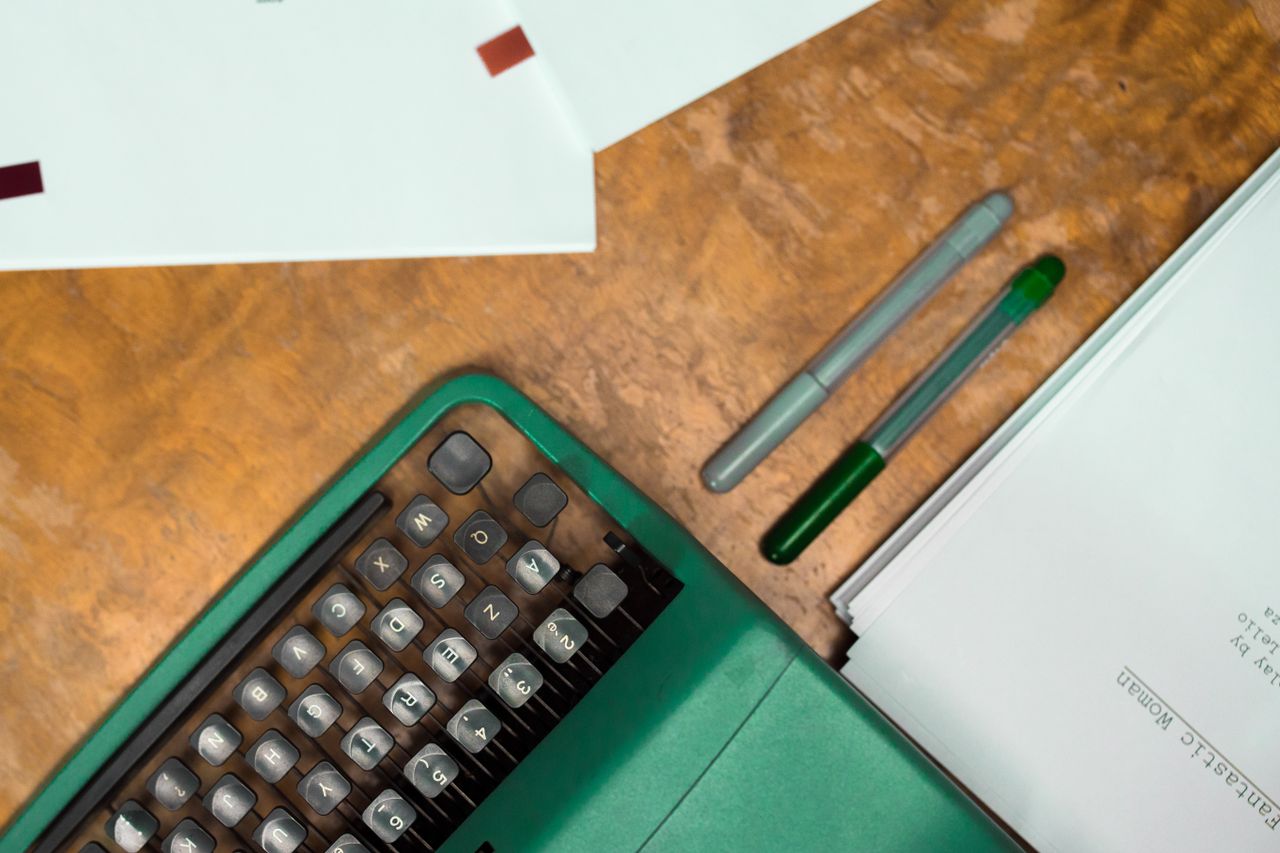 Flat lay of a green vintage typewriter, papers, and pens on a wooden desk.